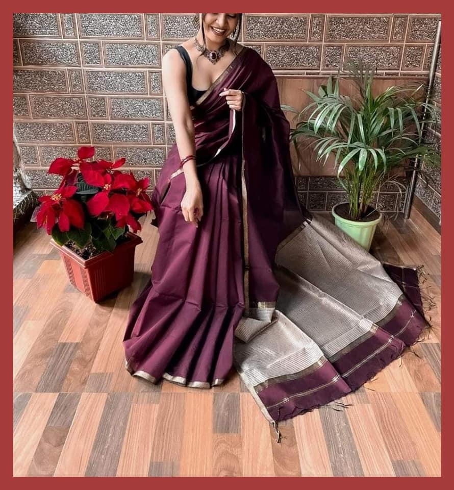 Model wearing a Mridula Maheshwari Saree in maroon with silver zari, seated indoors with indoor plants and red flowers in the background.
