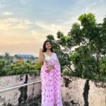 Woman draped in a soft pink cotton saree with red heart motifs and tassels, standing on a terrace at sunset surrounded by greenery.