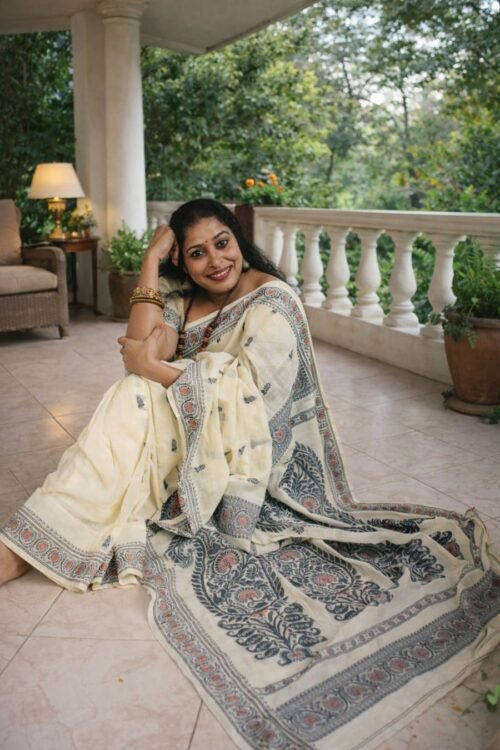 Indian woman seated on a veranda wearing an ivory cotton Baluchori saree with traditional woven motifs and ornate border